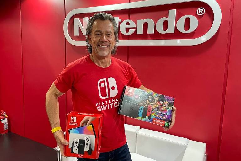 Um homem de vermelho, de meia idade, sorri enquanto segura consoles Nintendo Switch nas duas mãos. Ao fundo, o logo da Nintendo