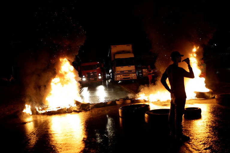 homem em frente de pneus em chamas com caminhões ao fundo 