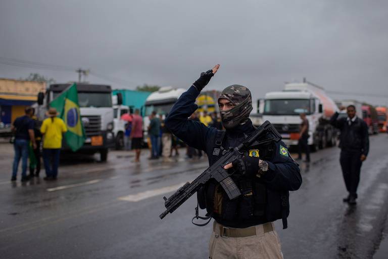 A imagem mostra um policial armado， usando uma máscara， em uma estrada onde há caminhões estacionados. O policial está levantando a mão em um gesto de sinalização. Ao fundo， há um grupo de pessoas， algumas com bandeiras do Brasil， e o céu está nublado， indicando um clima chuvoso.