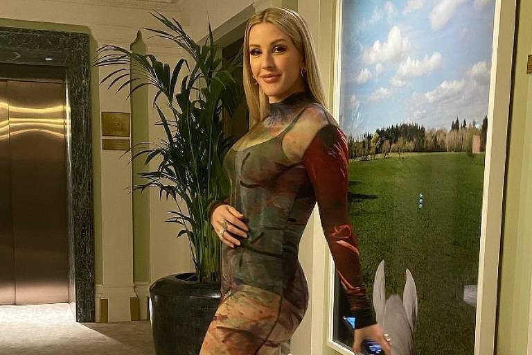 Em foto colorida, mulher usa vestido transparente e posa para foto no corredor de um hotel