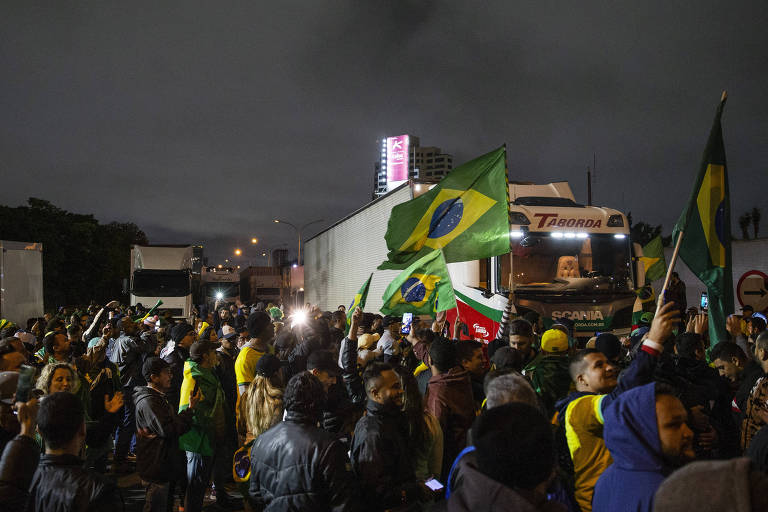 Foto feita à noite mostra várias pessoas com badeiras do Brasil ocupando um trecho de rodovia. Atrás delas, dois caminhões parados