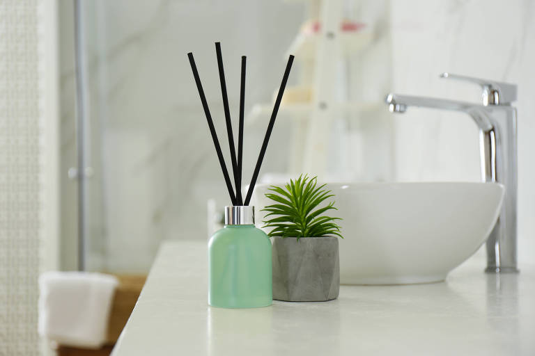 Sobre a bancada de um banheiro com pia branca, há um aromatizador de ambiente com varetas e um pequeno vaso de planta. 