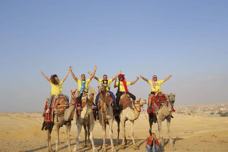 Cinco integrantes do Olodum estão de braços erguidos, cada um em um camelo, no Egito