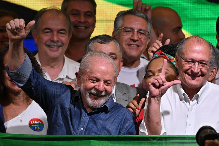 Lula é um homem idoso, branco, com poucos cabelos grisalhos. Ele está sorrindo e com as mãos levantadas, veste uma camisa social azul. Ele está cercado por uma multidão. Ao fundo é possível ver uma bandeira verde-amarela