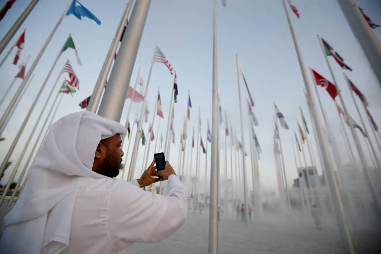 Pessoa com trajes árabes na cor branca registra em celular a Flag Plaza (praça das Bandeiras), uma das atrações turísticas de Doha, a capital do Qatar, país-sede da Copa do Mundo de 2022