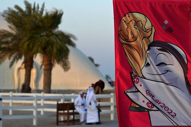 Banner com a imagem de La'eeb, mascote da Copa do Qatar, abraçada à Taça Fifa, enfeita praia de Doha; ao fundo, duas pessoas vestidas de branco e duas palmeiras