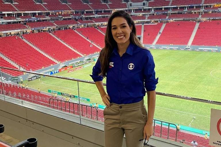 Em foto colorida, mulher de uniforme faz foto no meio de um estádio de futebol vazio