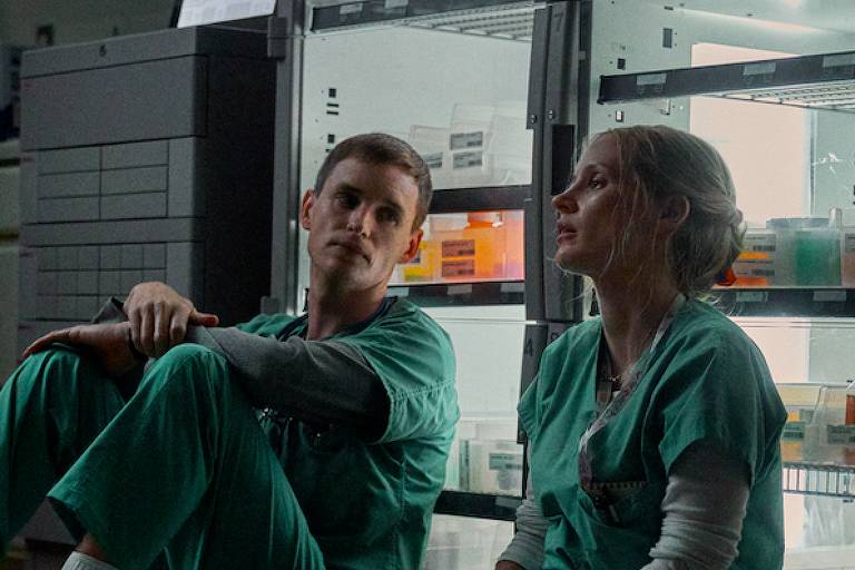 Fotografia colorida de Eddie Redmayne e Jessica Chastain sentados em ambiente hospitalar, de costas para um armário de medicamentos.