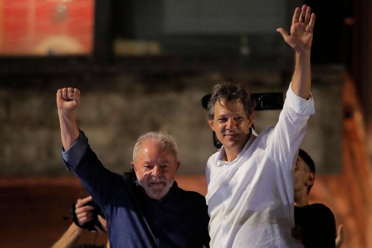 Lula, de camisa azul, levanta com sua mão esquerda a mão de direita de Haddad, de camisa branca, em frente a um fundo vermelho
