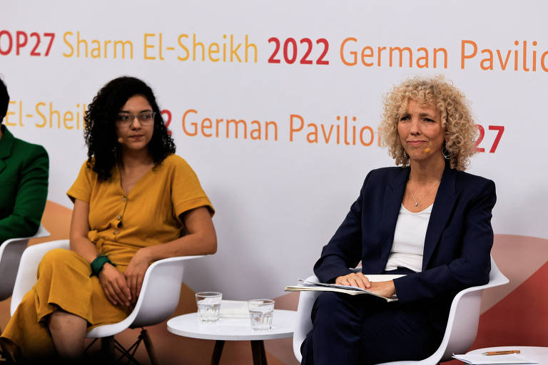Retrato de duas mulheres sentadas com painel ao fundo escrito com referências à COP27 e à Alemanha