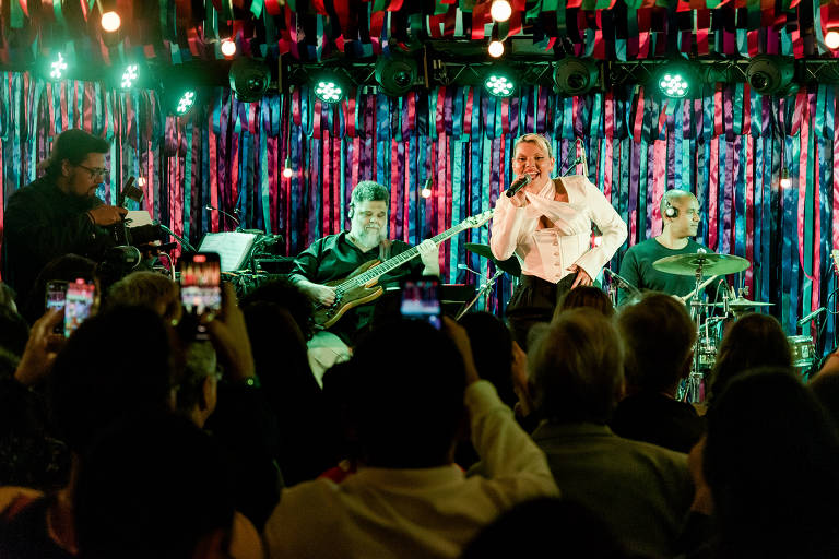 Em foto colorida, mulher de blusa branca canta em cima de um palco