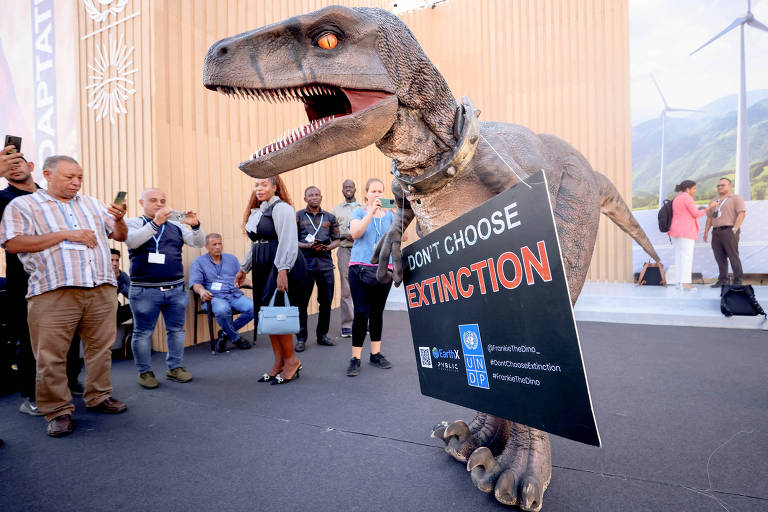 Pessoa fantasiada de dinossauro segura cartaz sobre extinção enquanto pessoas em volta observam