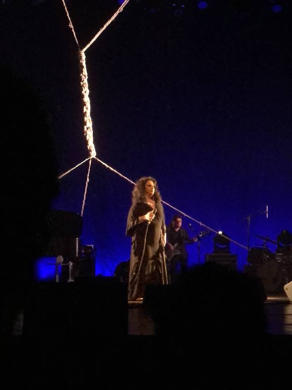 Gal Costa está em cima do palco, usando um vestido preto. o palco é iluminado por uma cor azul