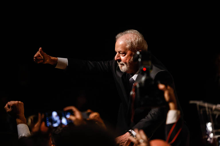 O presidente eleito Luiz Inácio Lula da Silva discursa durante reunião com parlamentares aliados. Ele é um homem branco, que veste um terno preto. Faz um sinal de positivo com a mão. 