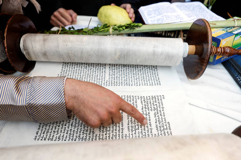 Imagem mostra braço e mão de um homem cujo dedo aponta para um rolo de texto da Torá, as Escrituras judaicas 