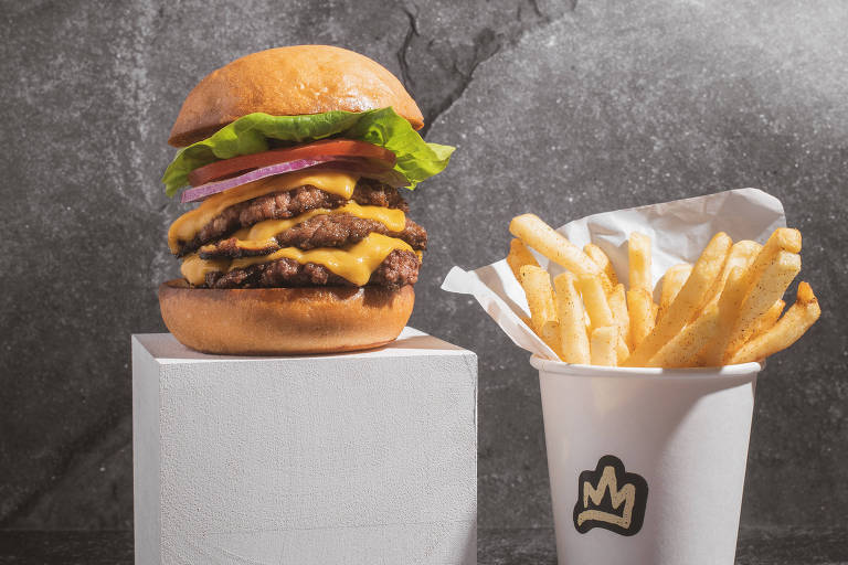 Com fundo cinza, foto mostra um hambúrguer que está em pé sobre um suporte quadrado branco; ao lado, um copo branco com porção de batata-frita