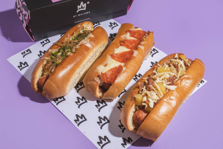 Foto mostra três hot-dogs dispostos sobre uma mesa lilás