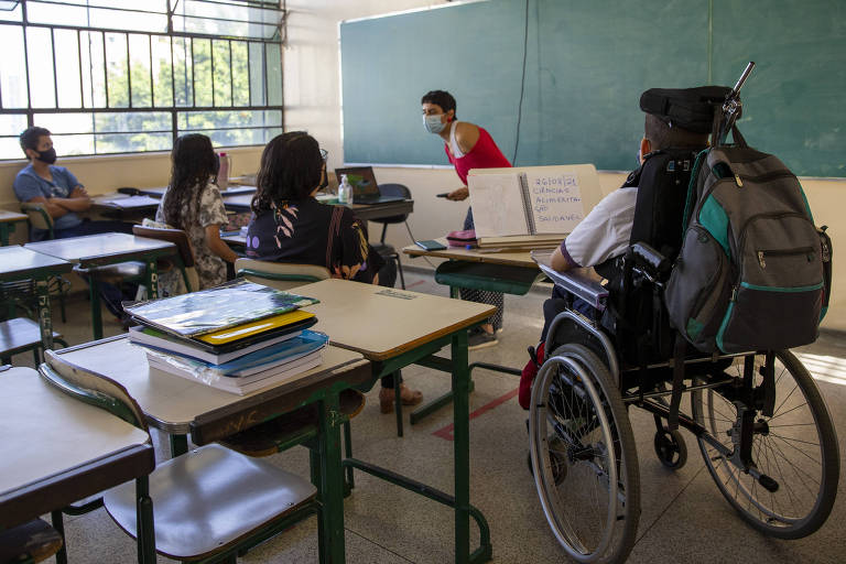 sala de aula com professora de pé, de vermelho, e alunos sentados. Um deles está de costas, em cadeira de rodas. 