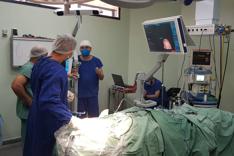 Imagem colorida mostra uma sala de cirurgia. No centro uma mesa cirúrgica, com um paciente coberto com um lençol verde. No lado esquerdo, dois médicos usam roupa azul e touca transparente. 