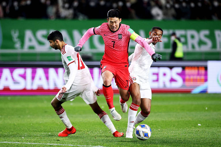 Heung-min Son, ao centro e com uniforme laranja, disputa bola com dois jogadores de uniforme branco, um de cada lado do atleta, em campo gramado de futebol