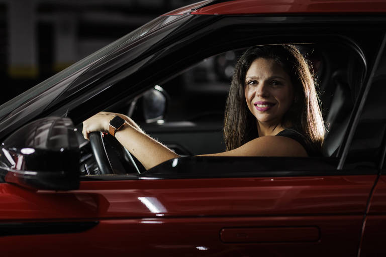 Moça de cabelos lisos e escuros olha para a câmera de dentro de um carro vermelho, com as duas mãos sobre o volante