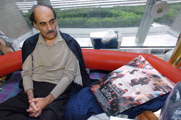 Mehran Karimi Nasseri, um homem calvo e de bigode, no canto do aeroporto de Paris onde morou por 18 anos; ao seu lado, o cartaz do filme de Steven Spielberg, inspirado em sua vida