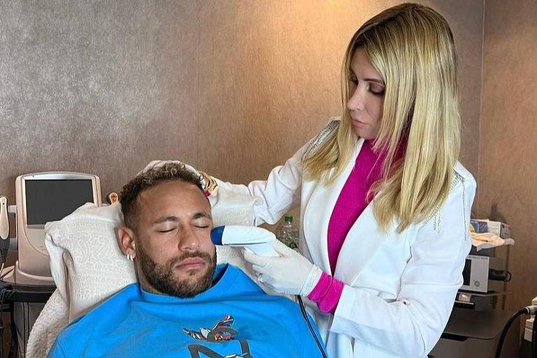 De casaco azul, Neymar está deitado na cadeira de um consultório enquanto a dermatologista passa um aparelho de laser em seu rosto