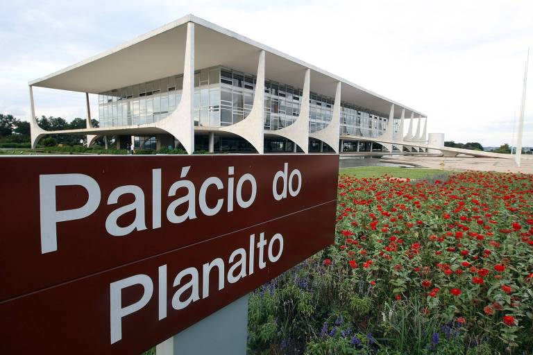 Prédio ao fundo. Na frente, uma placa com a frase "Palácio do Planalto". Próximo à placa, há flores no chão