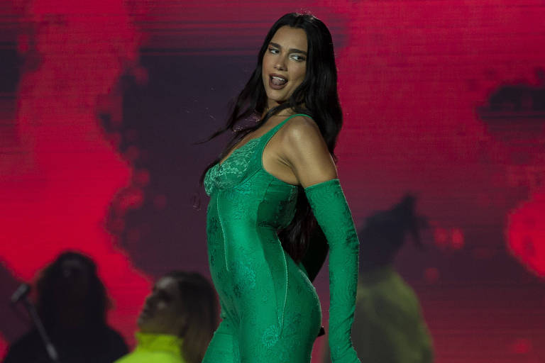 Em foto colorida,  mulher de macacão verde dança em cima do palco