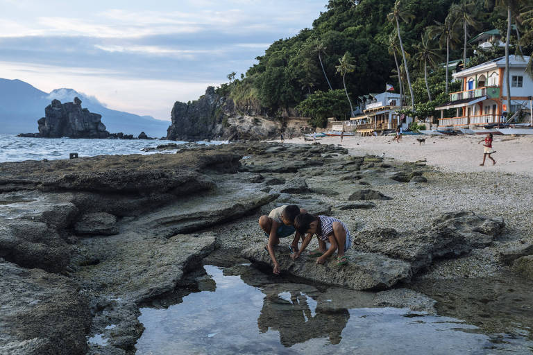 Crianças agachadas pegam iscas dentro de piscina natural formada por pedras à beira mar