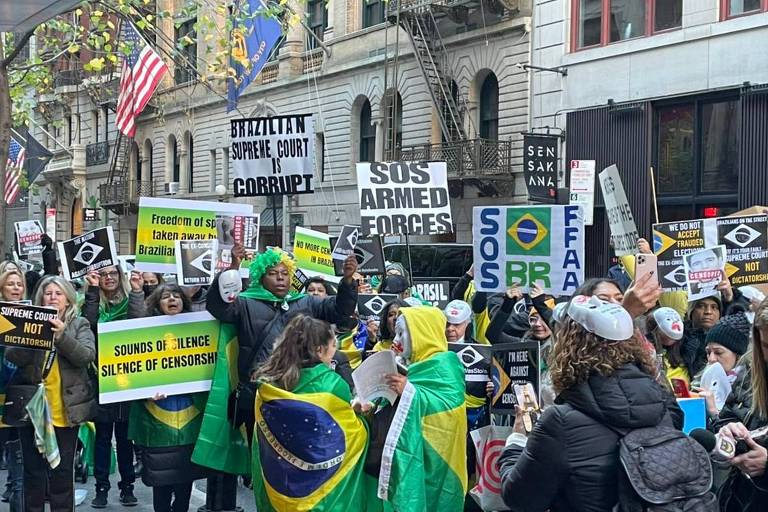 no meio de uma rua, em frente a um prédio com uma bandeira dos EUA, pessoas com cartazes em inglês e enroladas em bandeiras do Brasil