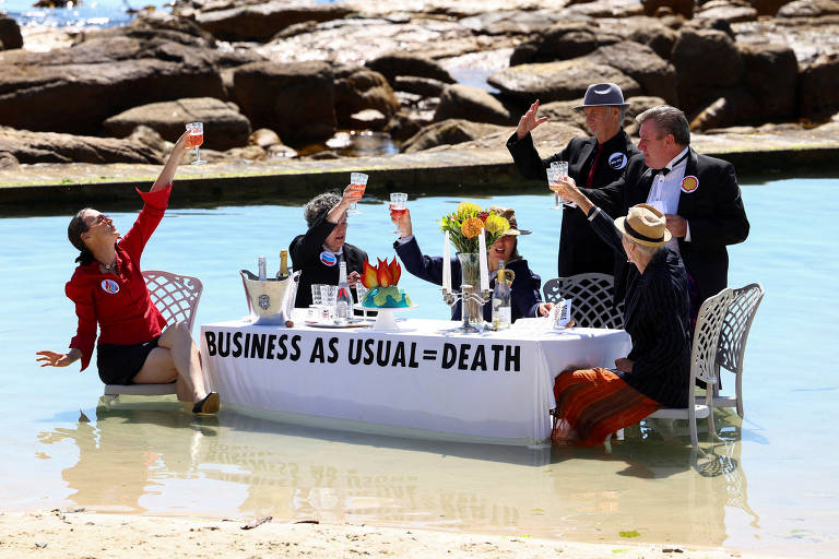 Grupo de homens e mulheres vestidos com roupas chiques brindam com drinques em volta de uma mesa de jantar posta dentro do mar à beira de uma praia