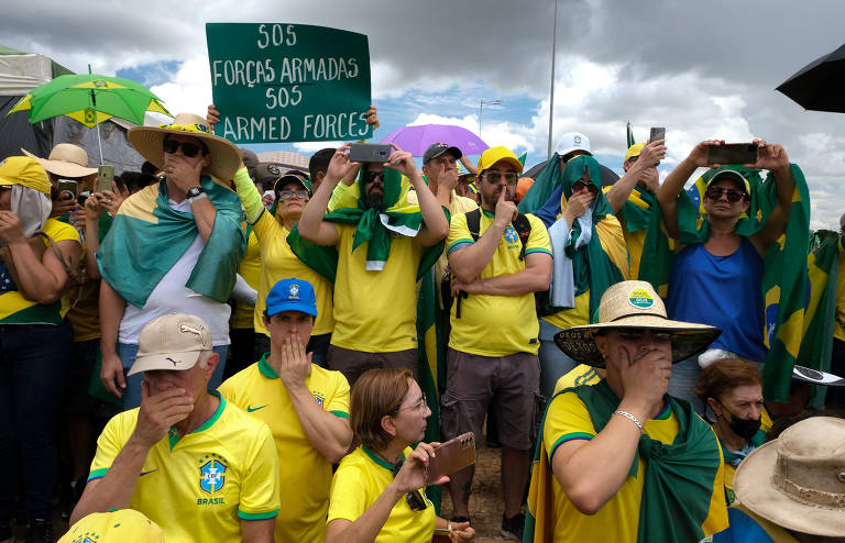 Foto mostra manifestantes bolsonaristas, com camisas da seleção brasileira, durante protesto em frente ao Quartel General do Exército, em Brasília. Um homem ergue um cartaz pedindo auxílio das Forças Armadas