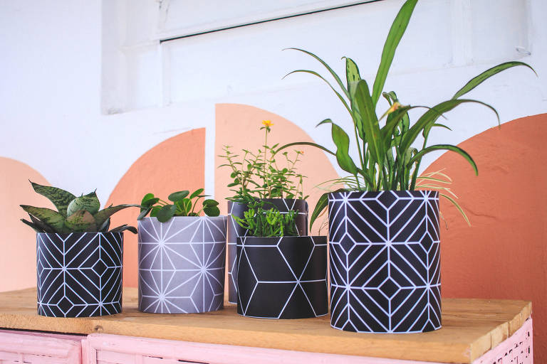 Vasos de plantas com desenhos geométricos em fundo preto ou cinza. Estão em cima de um armário de palha rosa e teto de madeira