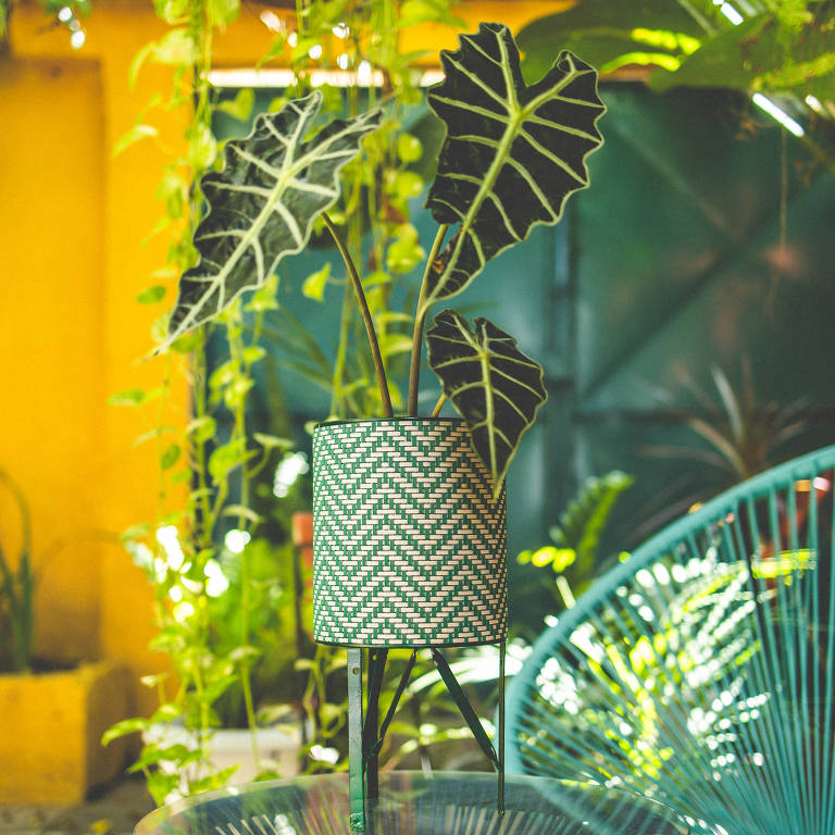 Vaso de planta feito com lata circular em verde e branco e três pés, que o deixam mais alto. O vaso está em cima de uma mesa azul.