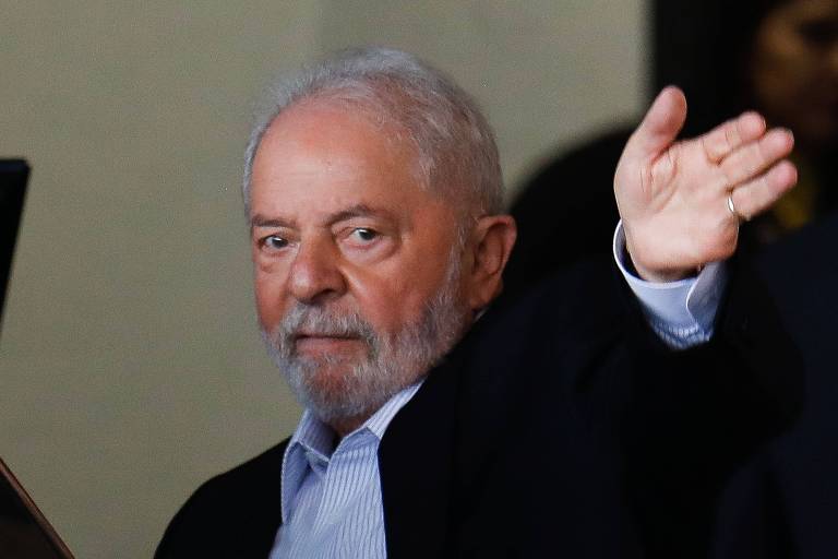 De camisa azul listrada e terno preto, |Lula estende o braço esquerdo, com a mão espalmada
