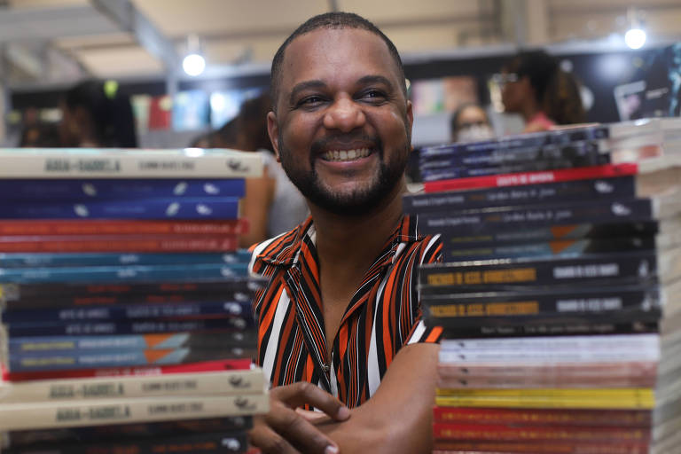 Retrato de Vagner Amaro, um homem negro com cabelo e barba curtos e pretos; ele sorri e veste uma camisa com listras verticais nas cores marrom, laranja, preto e branco; na frente dele, à esquerda e à direita, há duas pilhas de livros de sua editora, a Malê