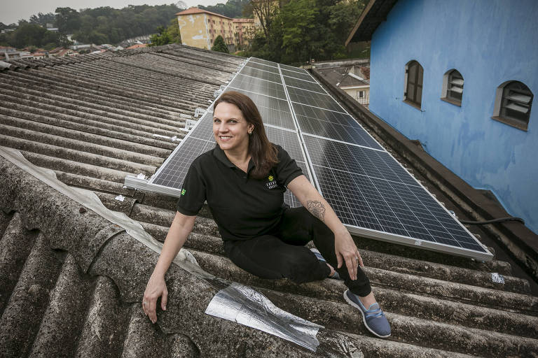 Imagem mostra mulher sentada em um telhado, onde há várias placas de painéis solares.
