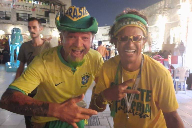 Sorridentes, lado a lado, usando chapéus nas cores verde e amarela e vestindo camisetas amarelas com estampas, os torcedores do Brasil Esdras Macedo, de 57 anos, e Wallace Leite, de 67 anos, posam para foto em Doha, no Qatar