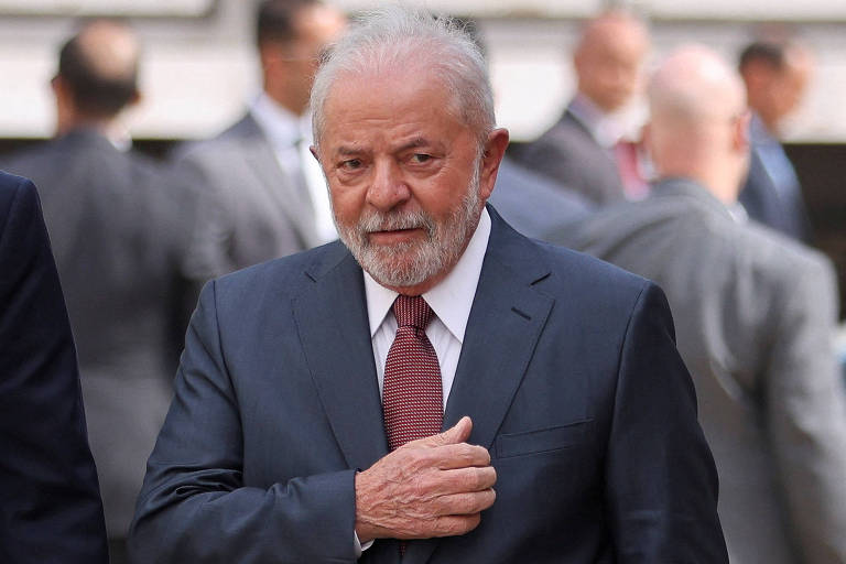 Fotografia colorida do presidente Lula em plano médio; ele veste um terno azul escuro, camisa branca e gravata cor de vinho e leva o braço direito dobrado na altura do peito, enquanto olha para a lateral