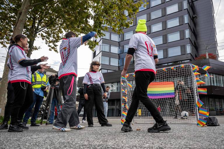 Pessoas de camiseta branca interagem em frente a uma trave de futebol montada na rua com a bandeira LGBTQIA+
