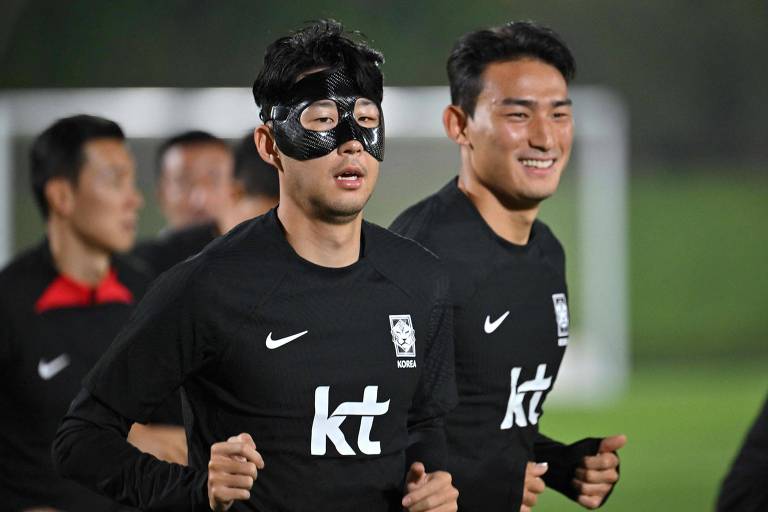 O atacante Son Heung-min, capitão da Coreia do Sul, treina com máscara de proteção, que cobre parcialmente seu rosto, em Doha, no Qatar; atrás dele, sorrindo, está seu colega de time Cho Yu-min