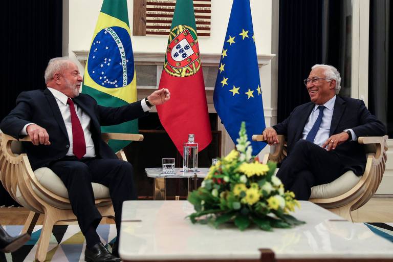 Lula viajou a Portugal em novembro, pouco após ser eleito, e foi recebido pelo primeiro-ministro luso, António Costa. Na imagem, os dois conversam sorridentes e sentados diante de bandeiras do Brasil, de Portugal e da União Europeia. São homens brancos e ambos estão de terno. Lula veste uma gravata vermelha e Costa, uma azul