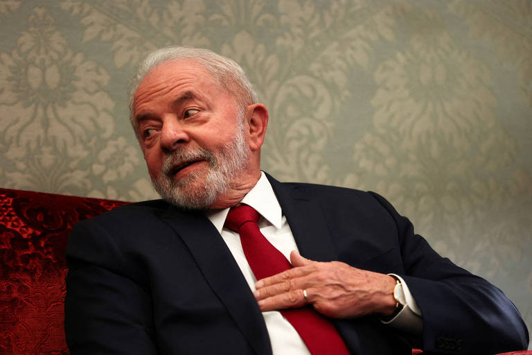 Lula, homem branco, tem cabelo e barba grisalhos. Está de camisa branca, gravata vermelha e paletó escuro, sentado em poltrona de tom vermelho. Está com a mão esquerda, que não tem o dedo mínimo, apoiada no peito