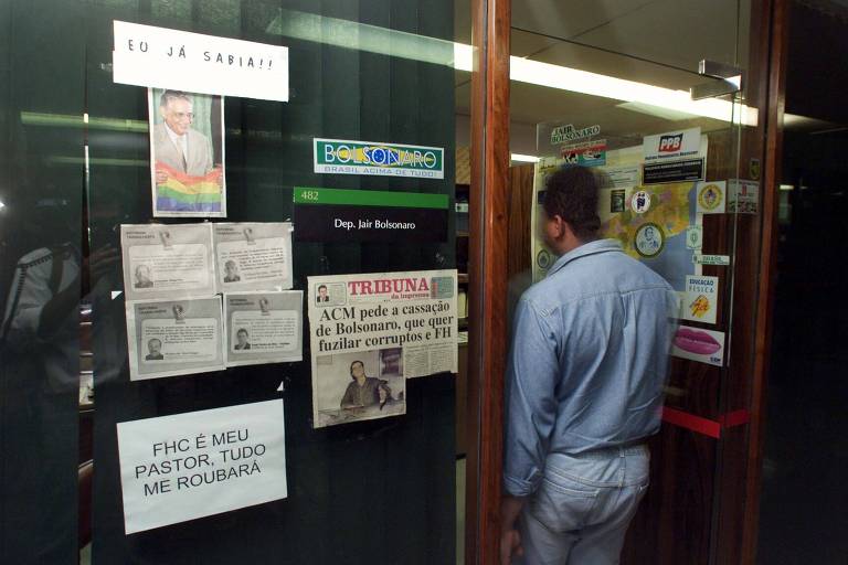 Fachada envidraçada do gabinete do então deputado Jair Bolsonaro, repleta de cartazes e adesivos. Em um, está escrito "FHC é meu pastor e tudo me roubará"