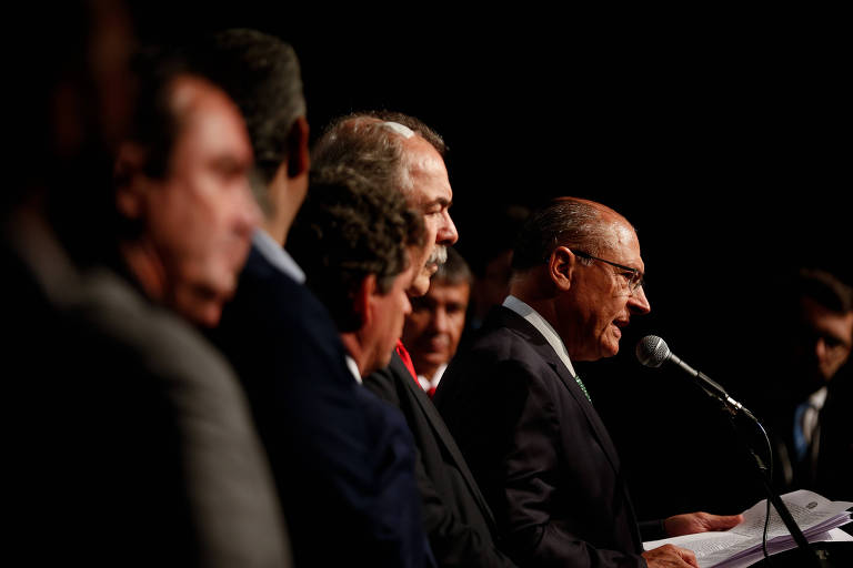 Foto mostra o vice-presidente eleito, Geraldo Alckmin, durante anúncio de novos nomes que vão integrar o governo de transição. Ele aparece ao fundo na imagem, lendo os nomes no microfone. É um homem branco, calvo e que usa óculos. Ele está ao lado de outros integrantes da equipe de transição