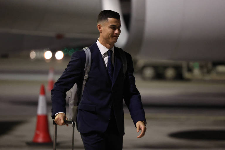 Puxando uma alça de mala com a mão direita e vestindo terno azul escuro, Cristiano Ronaldo caminha por área aberta do aeroporto depois do desembarque da seleção portuguesa