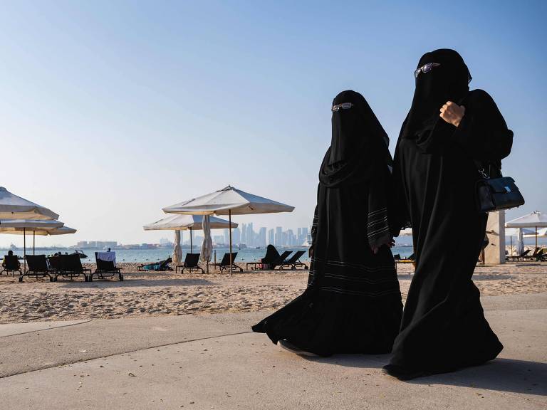 Vestidas de preto da cabeça aos pés, duas mulheres caminham em calçadão em Doha, em praia próxima ao estádio 974, um dos que abrigarão jogos da Copa do Mundo do Qatar; na areia há guarda-sóis abertos e, ao fundo, o mar