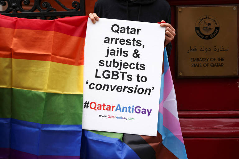Manifestante segura placa que encobre seus rosto e seu corpo com a inscrição 'Qatar detém, prende e sujeita LGBTs a conversão', durante ato próximo à embaixada do Qatar em Londres; ao lado há uma bandeira colorida que representa a causa LGBTQIA+