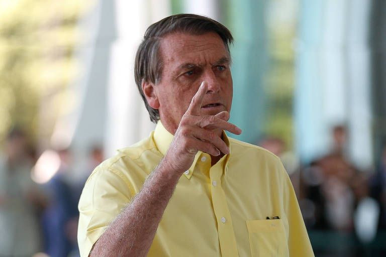 Na imagem, o presidente Jair Bolsonaro veste uma camisa amarela; ele está com o braço direito levantado até a altura do rosto